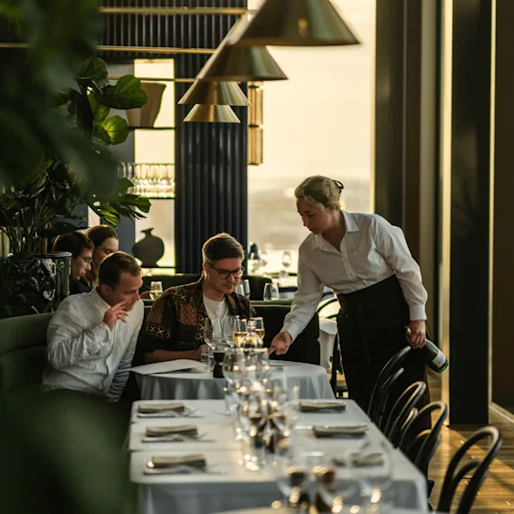 En tjener serverer gæster på en restaurant; elegant, velbelyst indretning med grønne planter.