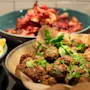 Sprøde kødboller pyntet med friske urter og ruccola fylder en skål, omgivet af tallerkener med røræg og sprødt bacon på en mørk køkkenbordplade.