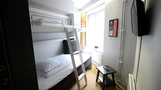 Kompakti hotellihuone, jossa on valkoinen kerrossänky tikkailla, pieni pöytä ja päivänvaloa tuova ikkuna.