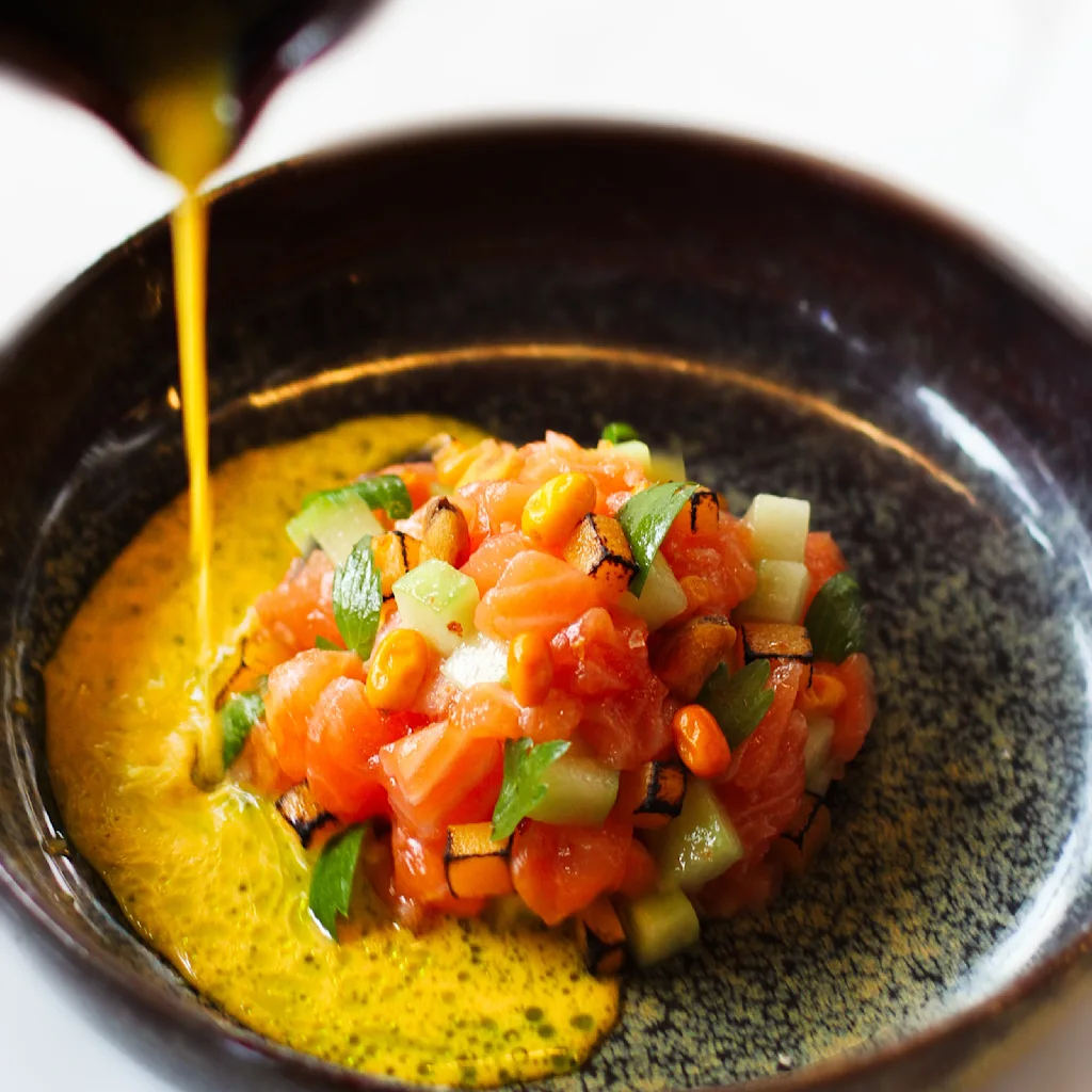 En bolle med terninger av laks, grønnsaker og urter serveres i en mørk bolle. En oransje-gul saus helles over, som fremhever de livlige fargene. Bakgrunnen er uklar.