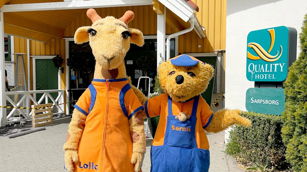 To maskoter, en sjiraff og en bjørn, står utenfor et Quality Hotel i Sarpsborg og ønsker gjester velkommen.