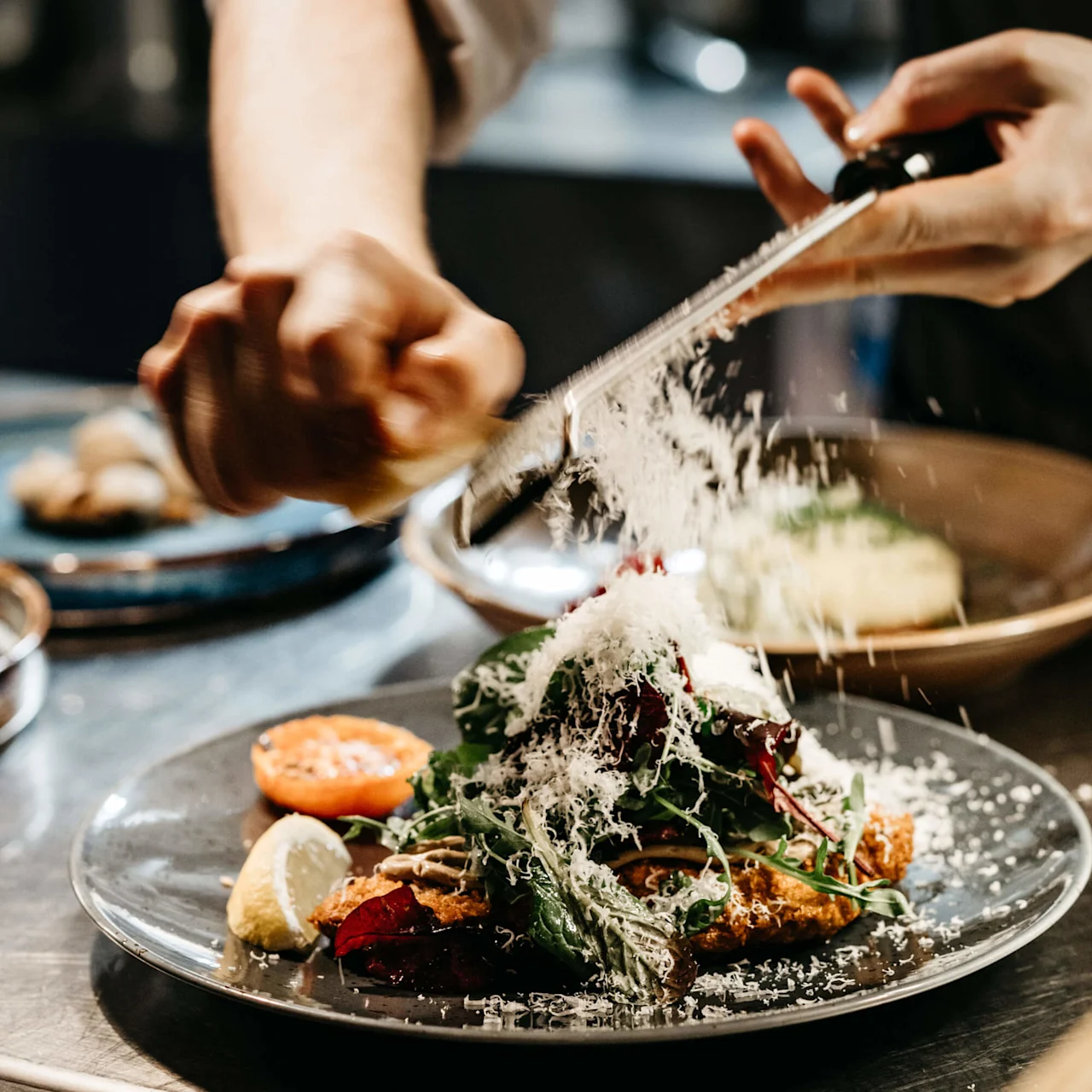 A chef grates cheese onto a salad in a bustling kitchen.
