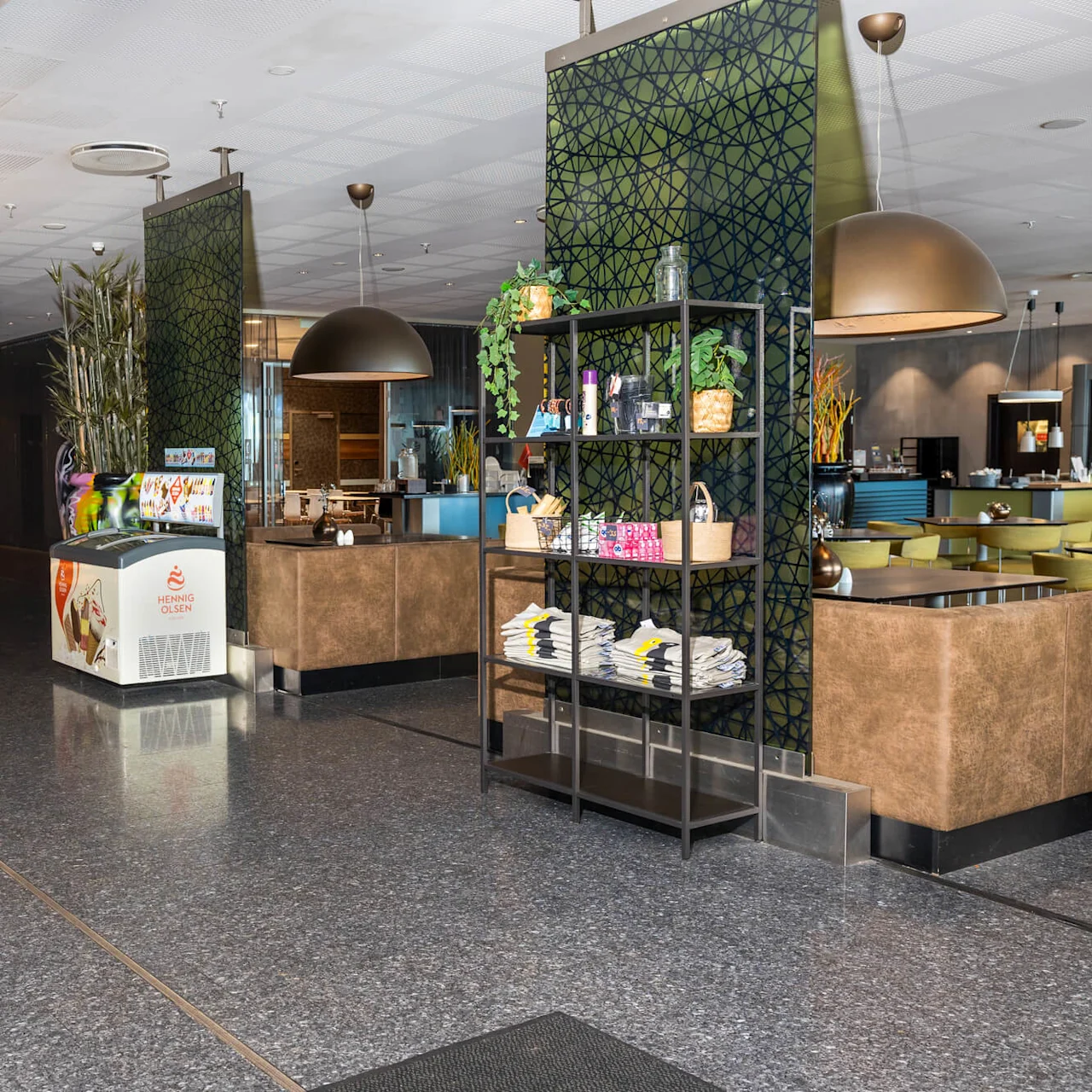 Reception desk with a man working, bar, and seating area surrounded by decorative panels in a hotel lobby. "Comfort Hotel Kristiansand" text above desk, ice cream freezer labeled "Hennig Olsen".