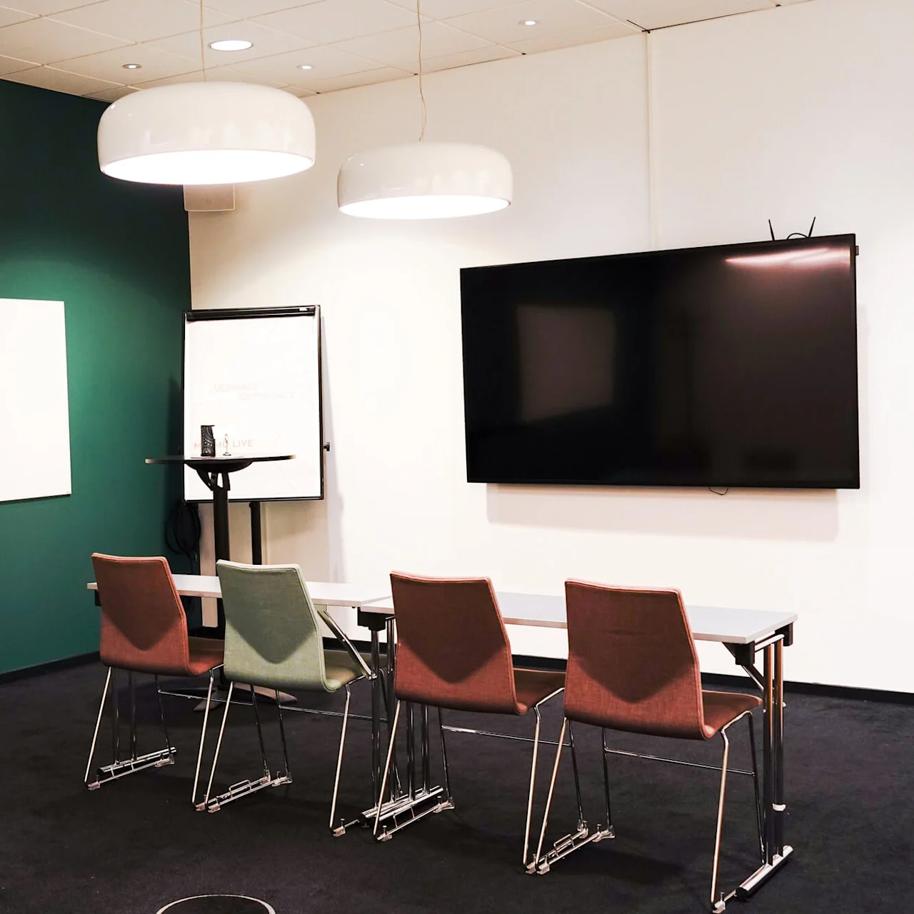 Et møterom med bord og stoler, grønn vegg med whiteboard, klokke og flippoverstativ. Stor TV-skjerm henger på tilstøtende vegg.
