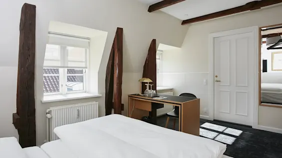 Et hyggeligt hotelværelse med en seng, et skrivebord og synlige træbjælker, med et vindue og en hvid dør.
