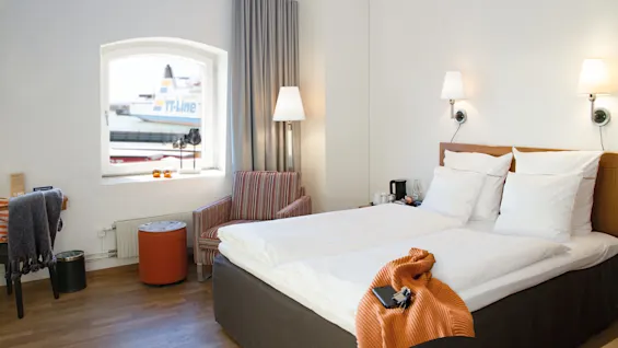 A hotel room with neatly made bed, a striped sofa, lamps, and a window showing a parked aircraft.
