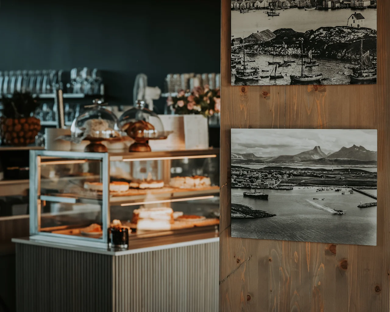 Et glasmontre indeholder et udvalg af bagværk under glaskupler. I nærheden pryder sort-hvide fotografier en trævæg. Miljøet ser ud til at være en hyggelig café.