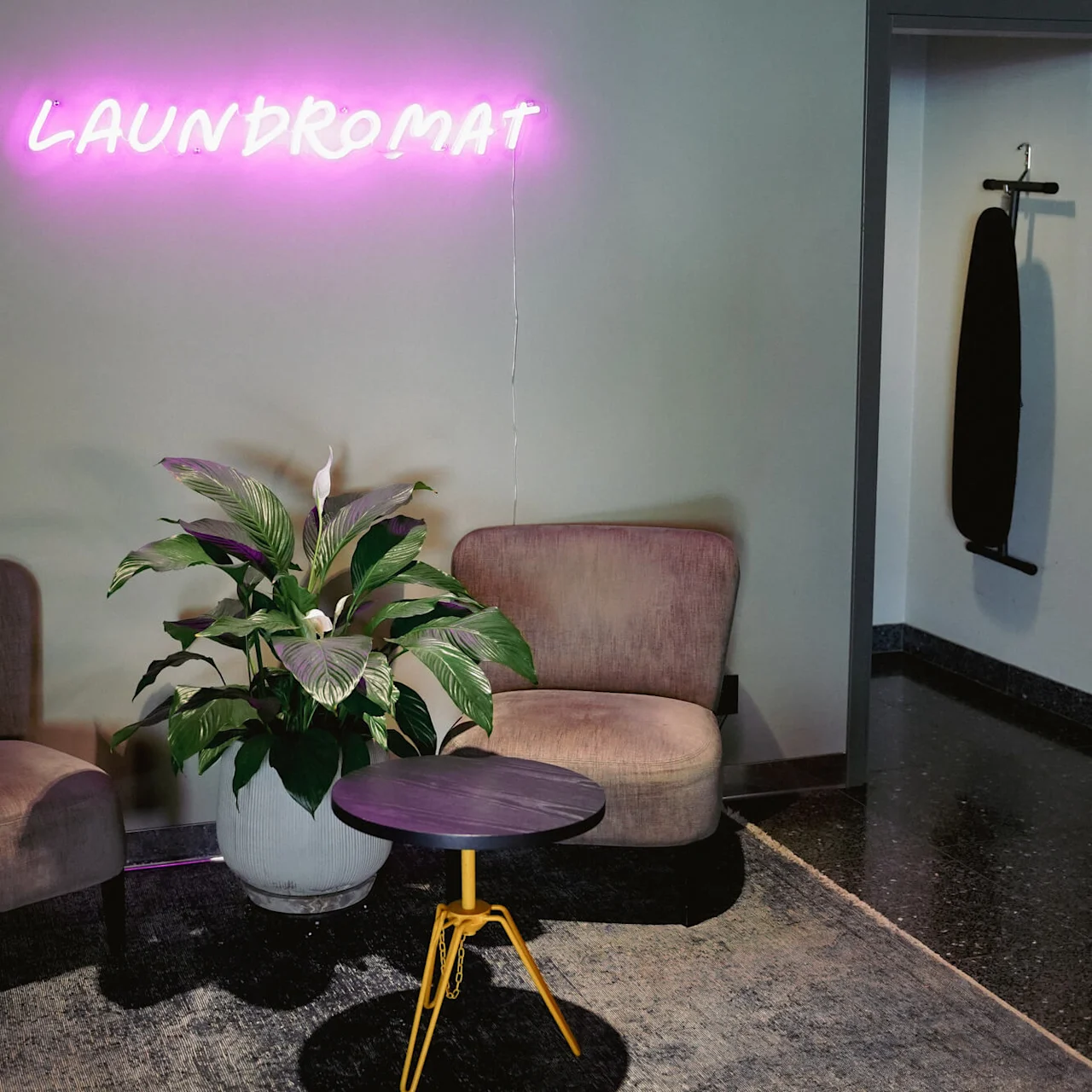 Two brown chairs and a plant next to a small table, under a neon sign reading "LAUNDROMAT" by a closed door with an ironing board.