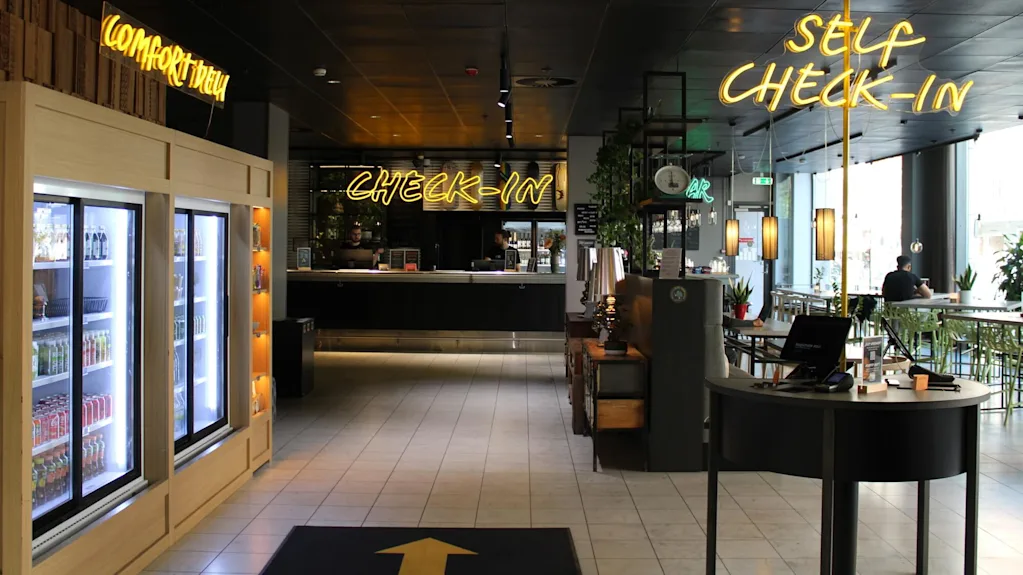 A modern hotel reception area with a self-service check-in station. Neon signs on the wall display the words "CHECK-IN" and "SELF CHECK-IN".
