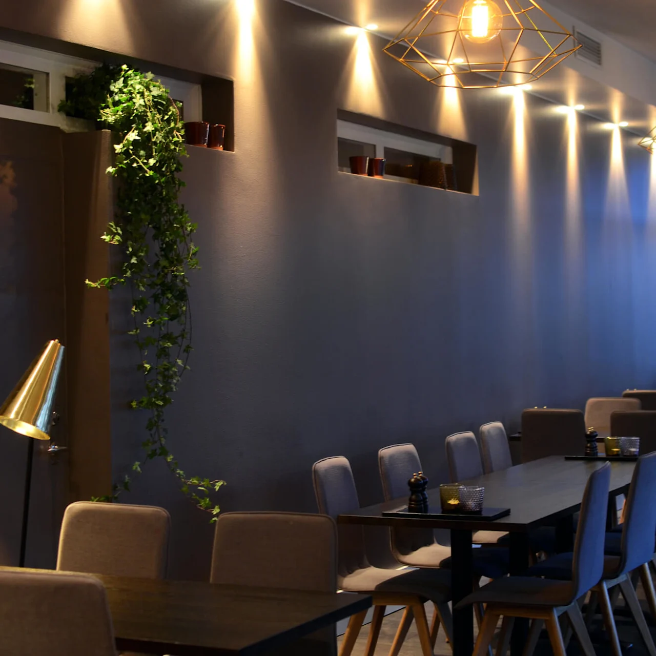 Modern restaurant interior with geometric hanging lights, upholstered chairs, and dark tables, highlighted by warm and cool lighting, creating a cozy ambiance.