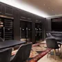 Dark dining tables and chairs are arranged in a restaurant setting. Wine coolers line the wall behind, with a mounted TV in the corner. A decorative carpet accents the floor.
