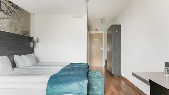 A bright and modern hotel room with two beds, a desk, and a wardrobe. An EXIT sign is visible above the door.