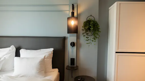 Et koselig soveromshjørne med en seng, hvitt sengetøy, en stilig nattbordslampe, et lite bord og en frodig hengeplante.