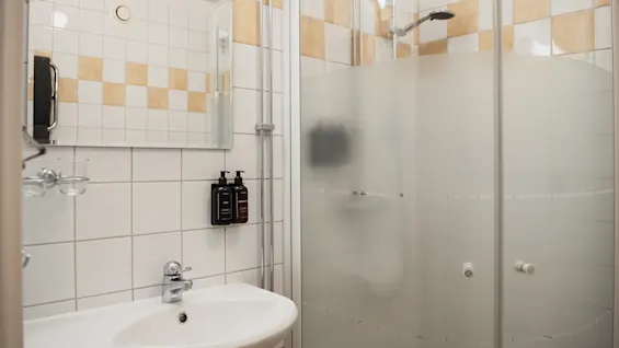 En hvit vask er montert på en flislagt baderomsvegg ved siden av en frostet glassdusj; såpedispensere er festet til veggen. Rommet har en moderne, minimalistisk design.