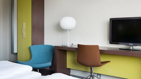 A modern room features a brown desk with a white lamp and a Philips TV. A green wardrobe, blue chair, and orange chair complete the minimalist design.