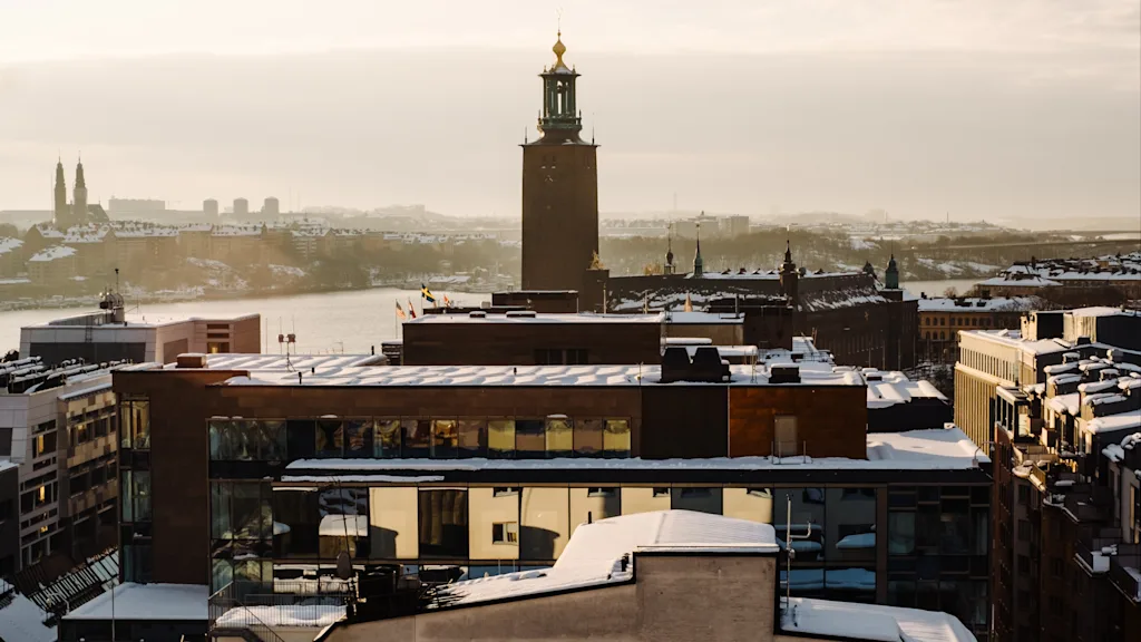 Norwegian Bokmål: Tak dekket av snø omgir en høy bygning med tårn. Bylandskapet ses ved solnedgang, med et varmt lys som faller over bylandskapet.