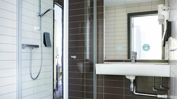 A modern bathroom features a glass shower with chrome fixtures and a white sink. Brown and white tiles surround, reflecting natural light. The text reads, “DRINK MORE WATER.”