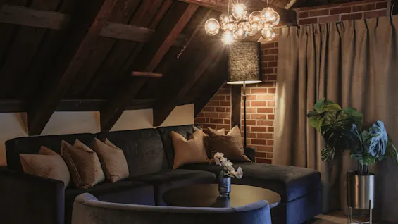 Et hyggeligt stueområde på et loft med komfortabel sofa, sofabord og varm belysning, træbjælker og murstensvæg.
