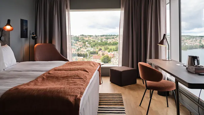 Hotellrom viser en pent oppredd seng, brunt teppe og lenestol. Skrivebord med lampe og vannkoker vender mot store vinduer med byutsikt under spredte skyer.