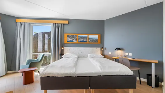 A modern bedroom with twin beds, a desk, chair, and artwork, illuminated by natural light from a window.