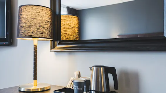 En tændt bordlampe og en elkedel med kopper mærket "hug in a mug" står på en bakke under et spejl på et skrivebord i et rum.