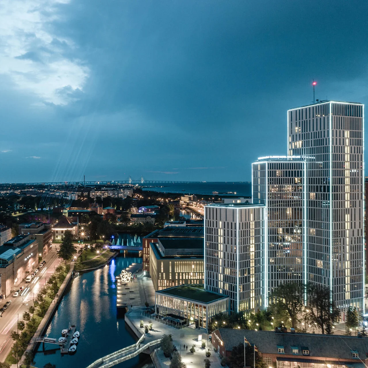 En høy, moderne skyskraper med opplyste konturer står ved siden av en kanal med fortøyde båter, i et livlig bymiljø under en skyet kveldshimmel.