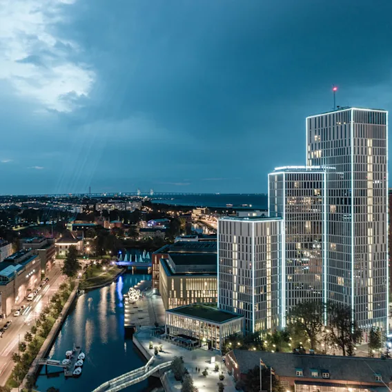 En hög, modern skyskrapa med upplysta konturer står bredvid en kanal med förtöjda båtar, i en livlig stadsmiljö under en molnig kvällshimmel.
