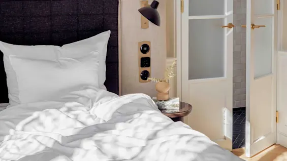 Bed with white pillows and sheets is next to a bedside table holding a small vase and book. A partially open door with glass panels reveals a tiled area.