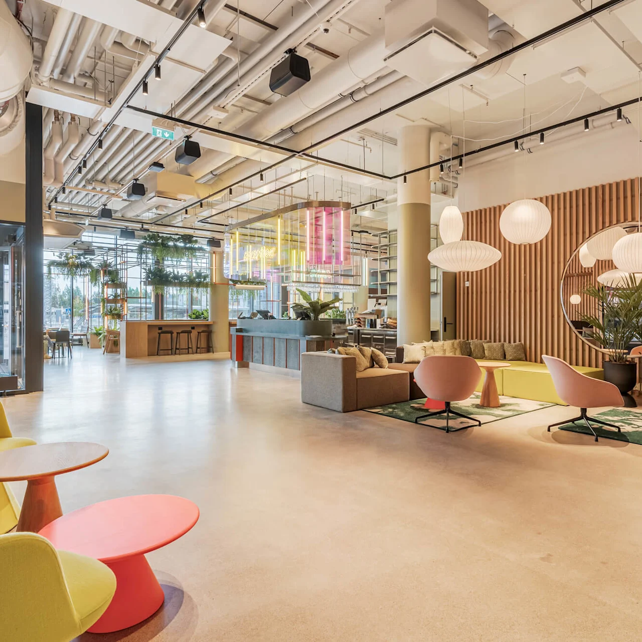 Lys, moderne lounge med farverige stole og borde arrangeret afslappet. Overhængende pendellamper oplyser træpanelvægge pyntet med planter. Neonskilt tilføjer liv i et luftigt, rummeligt miljø.
