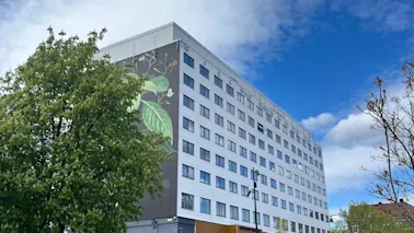 Byggnad med lövmålning står vid trädkantad gata under delvis molnig blå himmel. Bilar och fotgängare syns.