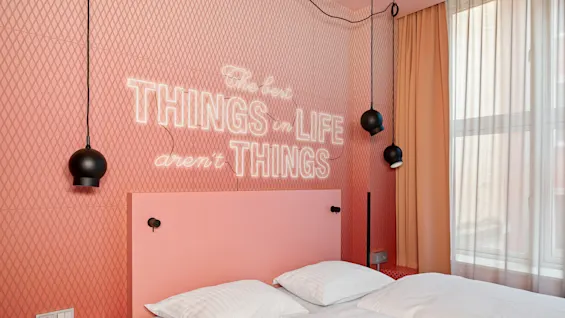 En seng med hvitt sengetøy mot en rosa vegg med teksten: "The best THINGS in LIFE aren't THINGS" i lysende bokstaver, omgitt av svarte pendellamper.