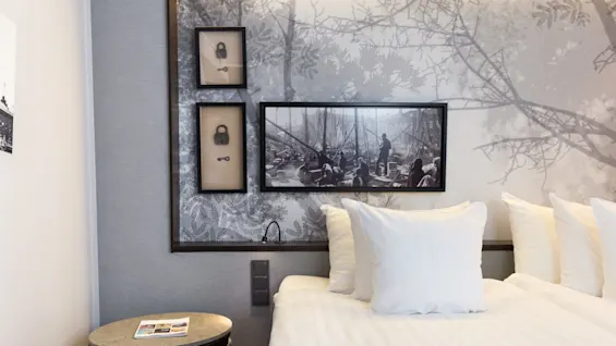 A neatly-made bed with white linens in a room, decorated with black-and-white framed images on the wall, alongside a patterned wallpaper.