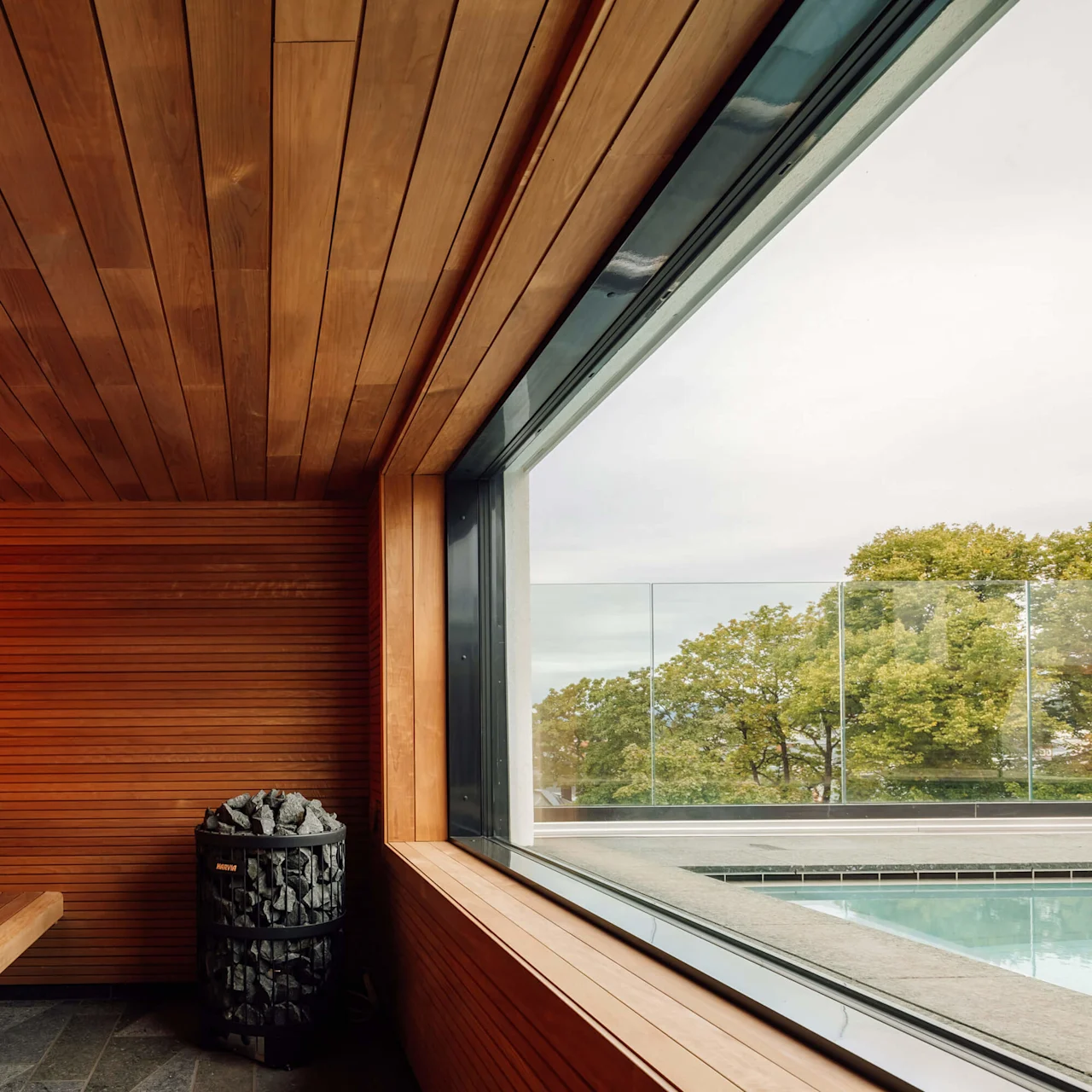 Træbeklædt sauna interiør med komfur, bænksæder, og glasvægge der giver udsigt til træer og en pool.