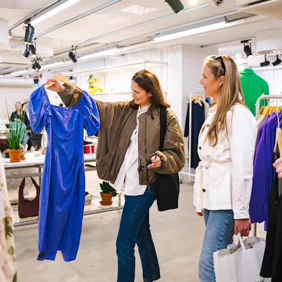 Tre kvinnor shoppar i en klädbutik, en håller upp en blå klänning för sina vänner att se.