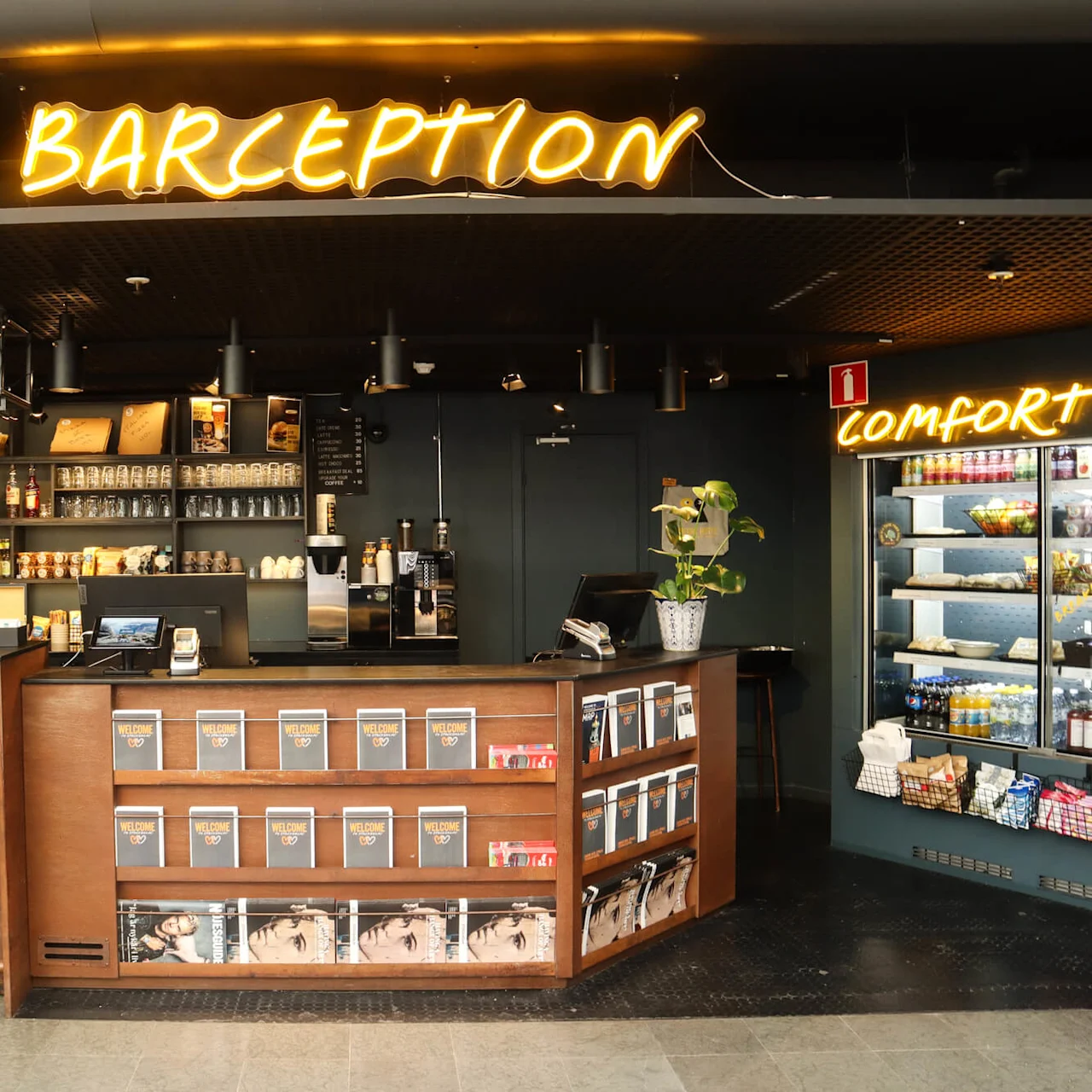En bar med belyst "BARCEPTION"-skilt over og et "COMFORT B"-skilt til højre. Magasinstativ og et køleskab med snacks er en del af omgivelserne.