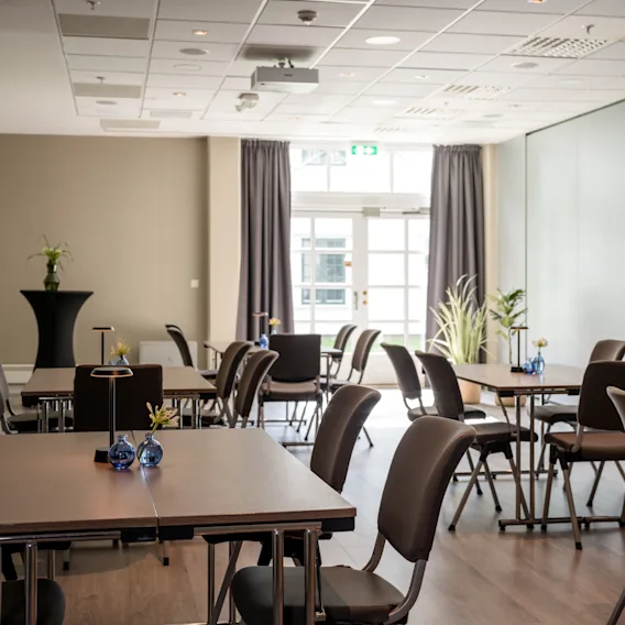 A modern, bright conference room with multiple tables and chairs arranged for a meeting, featuring a projector and large windows.