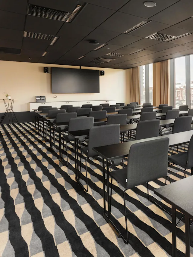 Rader av svarta stolar och bord är uppradade i ett modernt konferensrum med en stor skärm. Rummet har en mönstrad matta och stora fönster med utsikt över en stadsbild.
