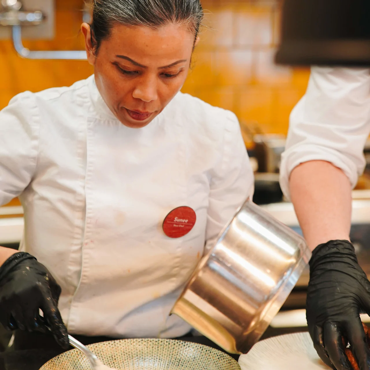 Kok i en hvid uniform fokuserer på at anrette mad fra en gryde på en tallerken i et professionelt køkken. Skiltet siger: "Sumee, Head Chef."