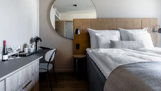 A neatly made bed with white pillows and a gray blanket sits beside a desk with a chair, a lamp, wine, glasses, and a coffee set, in a modern room.