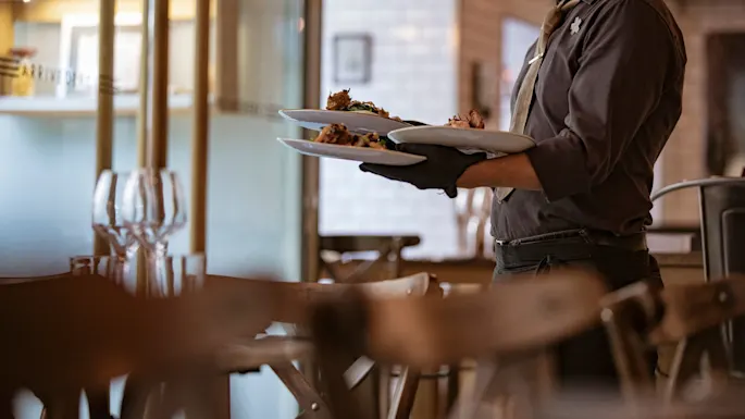 En servitør med mørke hansker bærer flere tallerkener med retter gjennom en moderne restaurant som er varmt belyst, med trebord og stoler. En hylle med svakt opplyste gjenstander vises i bakgrunnen.