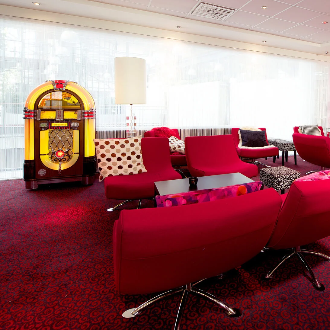 A vibrant lounge with red chairs, a classic jukebox on the left, and a large, colorful wall art piece in a modern setting.
