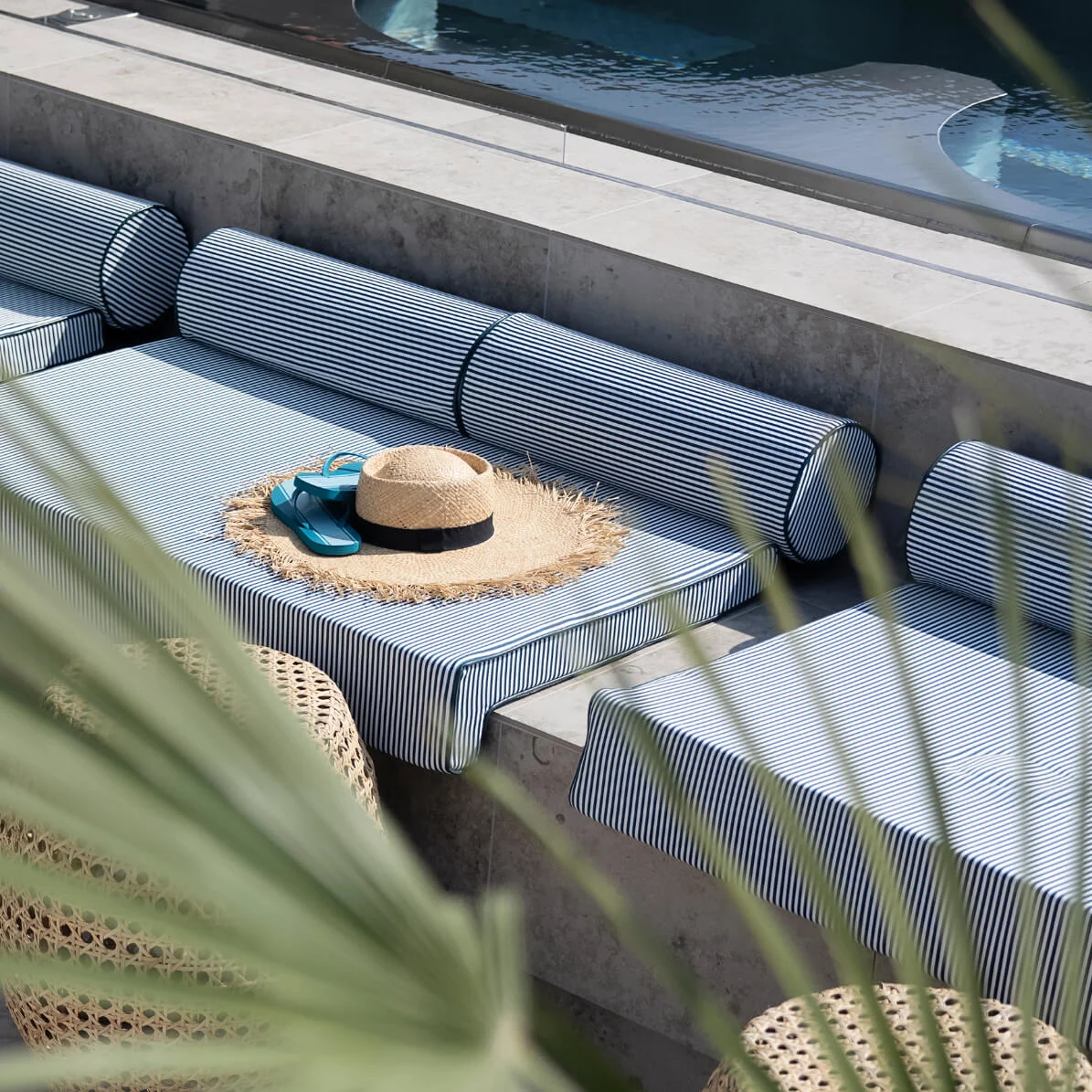 Striped cushions rest on a concrete ledge beside a pool, with a straw hat and blue flip-flops placed on them. Wicker stools and palm leaves surround the scene.