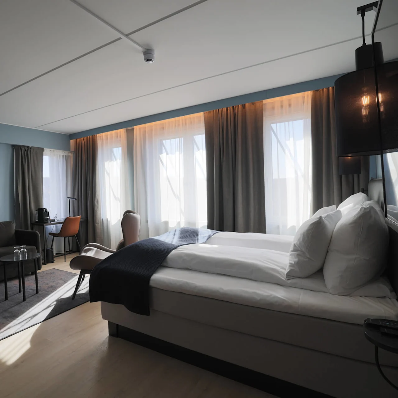 A well-lit, modern hotel room with a bed, sofa, desk, and large windows with sheer and opaque curtains.