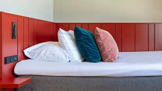 A red bed headboard, white bedding, and colorful pillows in a room with plain wall.