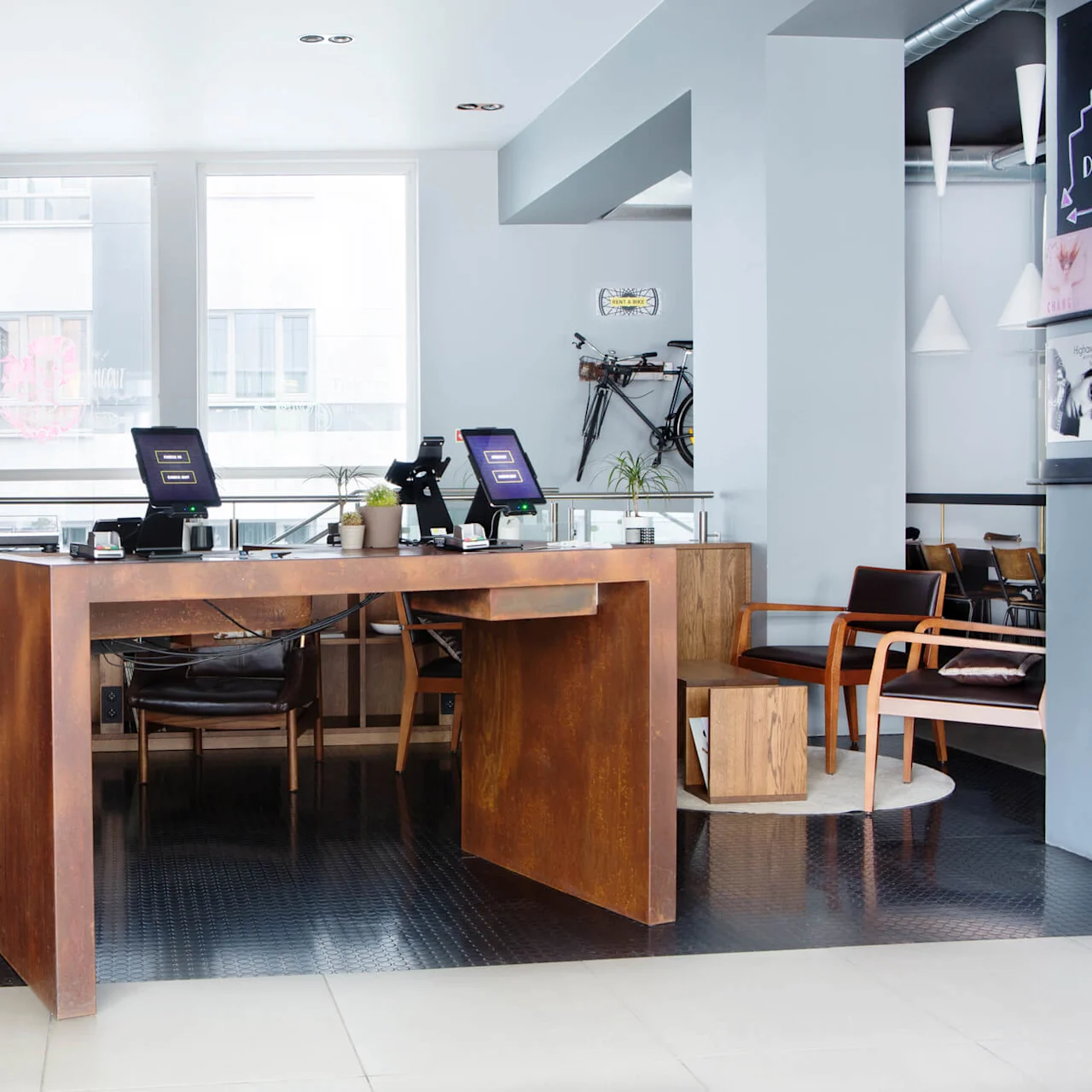 Modern wooden desk with two computers displaying screensavers, surrounded by brown chairs in a bright office with large windows, a colorful map, and a bicycle on the wall.