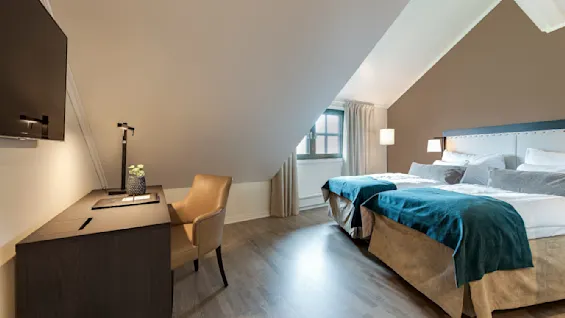 Et ryddig soverom med en stor seng dekket med hvitt og blått sengetøy ligger under et skråtak. Et skrivebord med en lampe og stol står i nærheten.