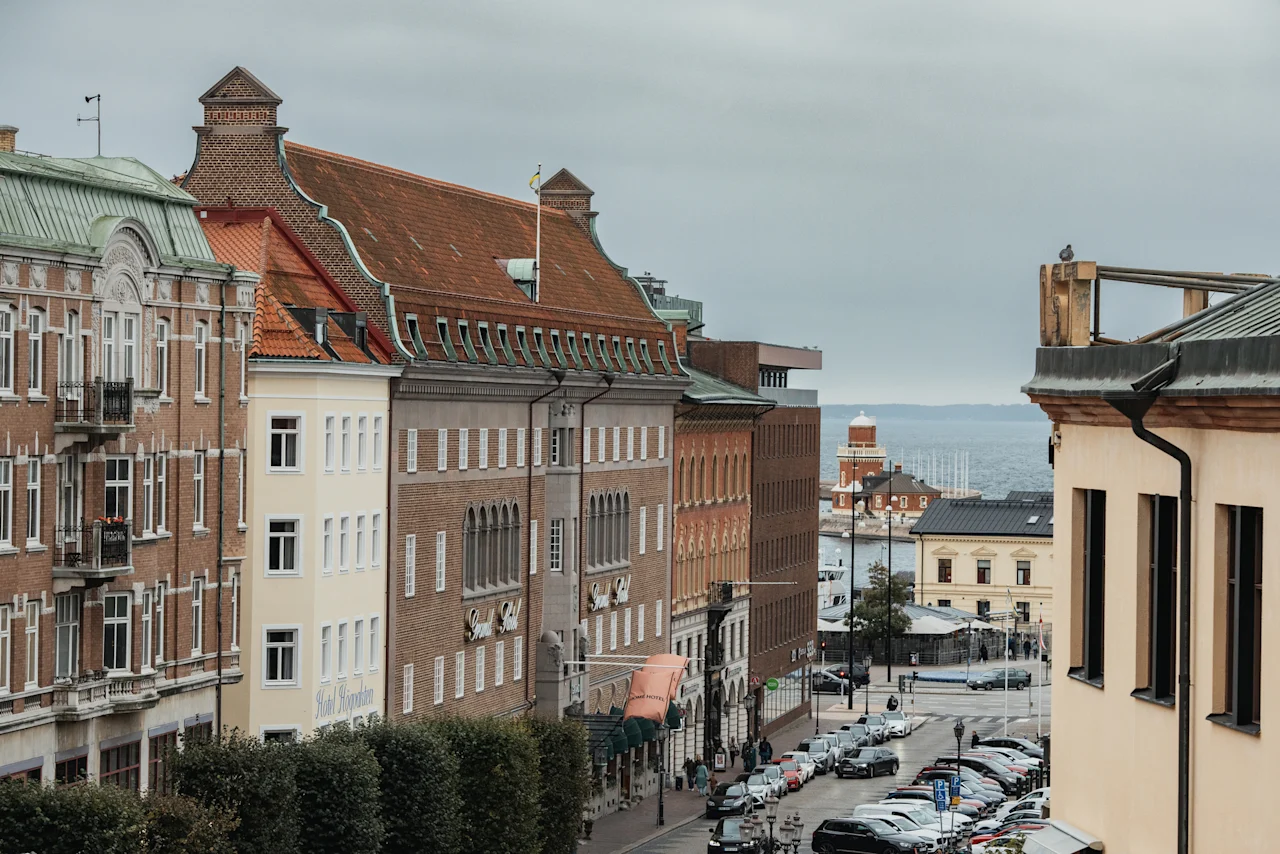 En gatuvy i Helsingborg med historiska byggnader, parkerade bilar och en glimt av havet och en fyr. Hotel Grand är synligt.