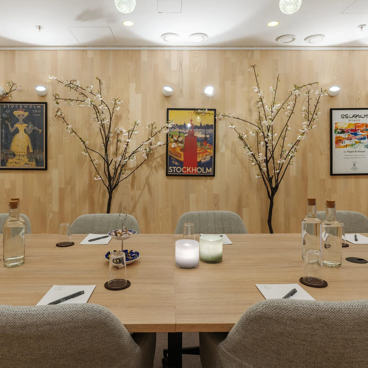 A wooden table holds glass bottles, notepads, and pens beneath a wall adorned with three framed "Stockholm" posters and decorative branches with blossoms, creating a serene meeting atmosphere.