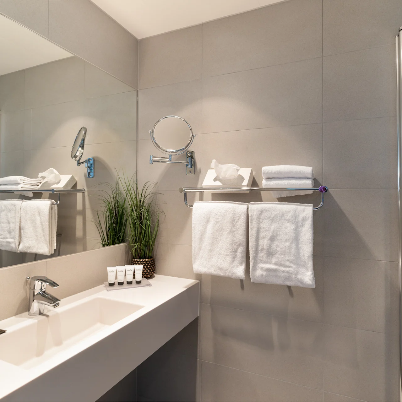 A modern bathroom features a sink with toiletries, adjacent towels on a rack, and a wall-mounted mirror. The setting is sleek with gray tiled walls.