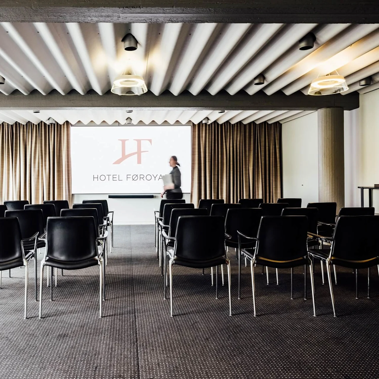 Rækker af tomme sorte stole vender mod en præsentationsskærm med teksten "HOTEL FØROYAR." En person går over det tæppebelagte konferencelokale omgivet af gardiner og lys.